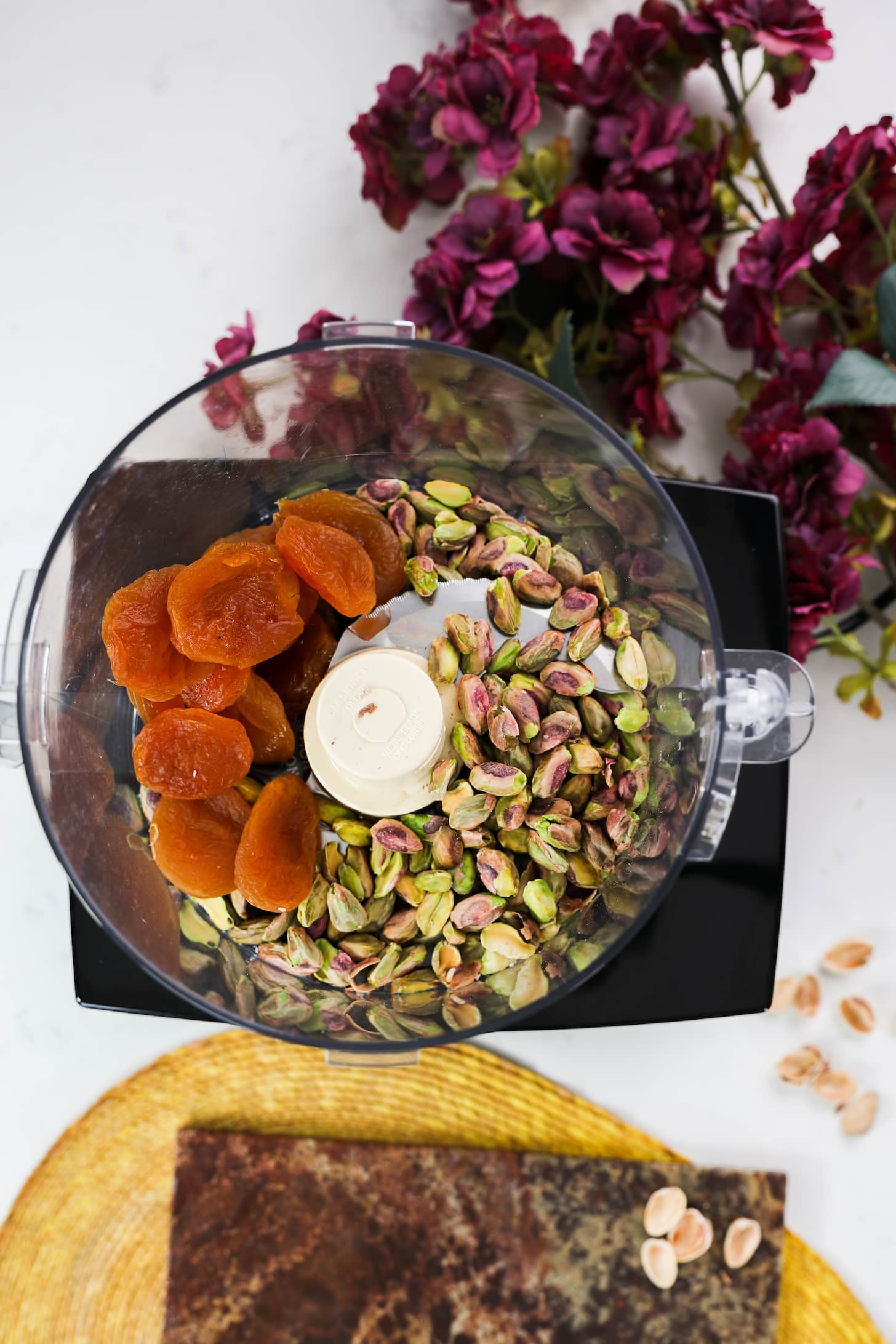 food processor containing pistachios and dried apricots with flowers on one side of a worktop surface.
