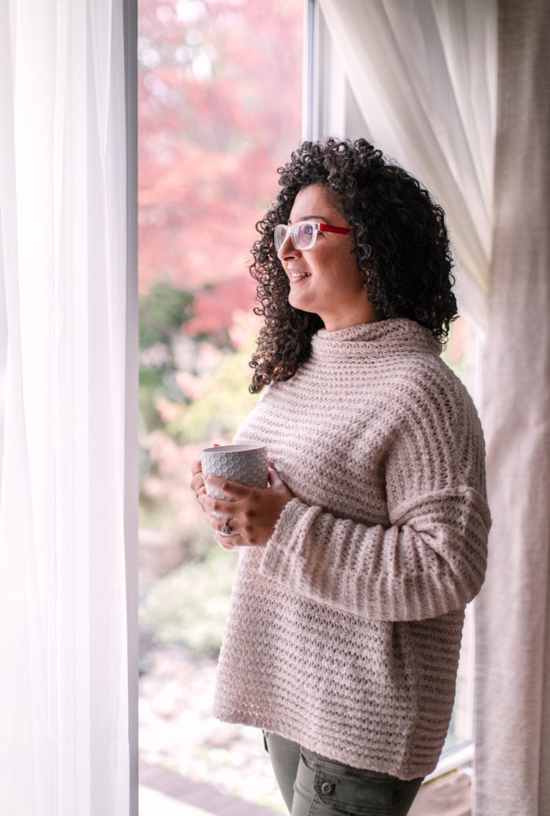 curly haired lady looking out of a window holding a mug.