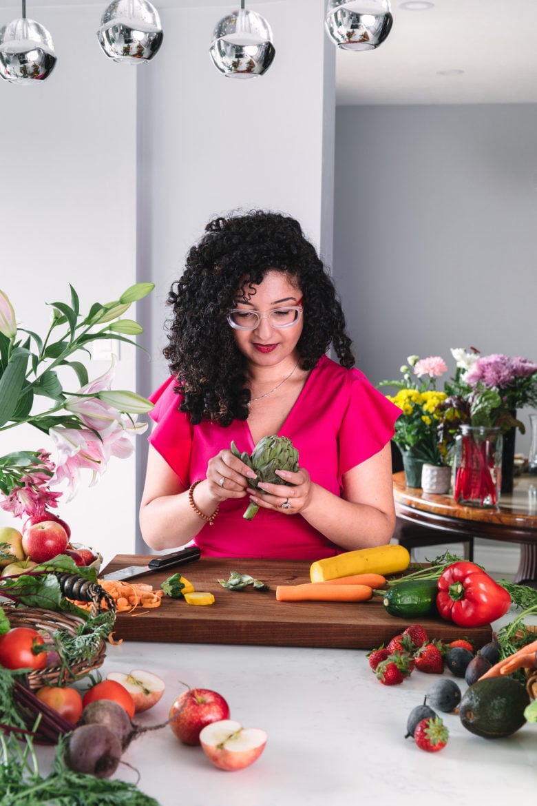a lady standing behind a worktop peeling an artichoke surrounded by colourful produce and flowers.