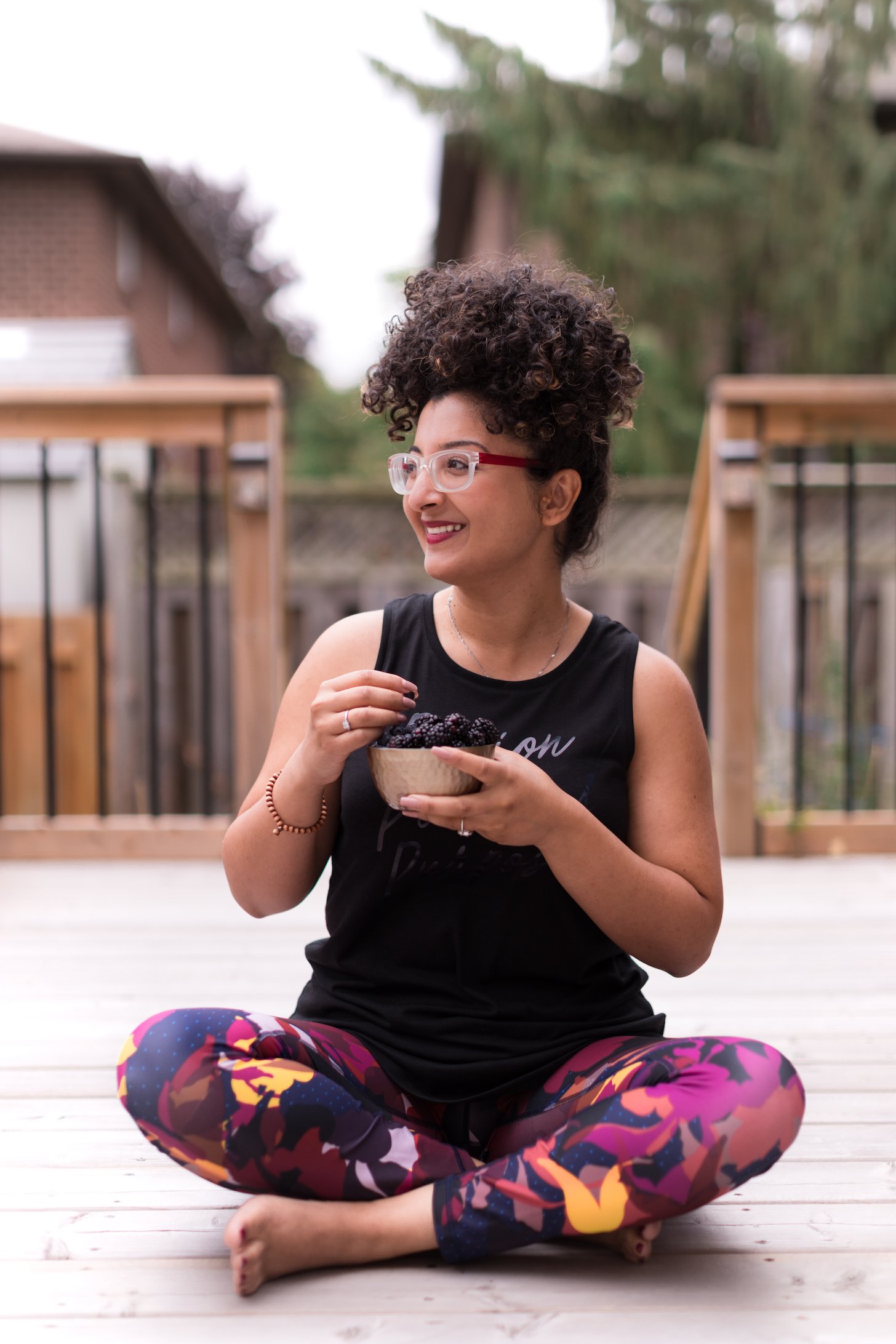 a lady sitting in a yoga position on a wooden deck outside holding a bowl of berries.