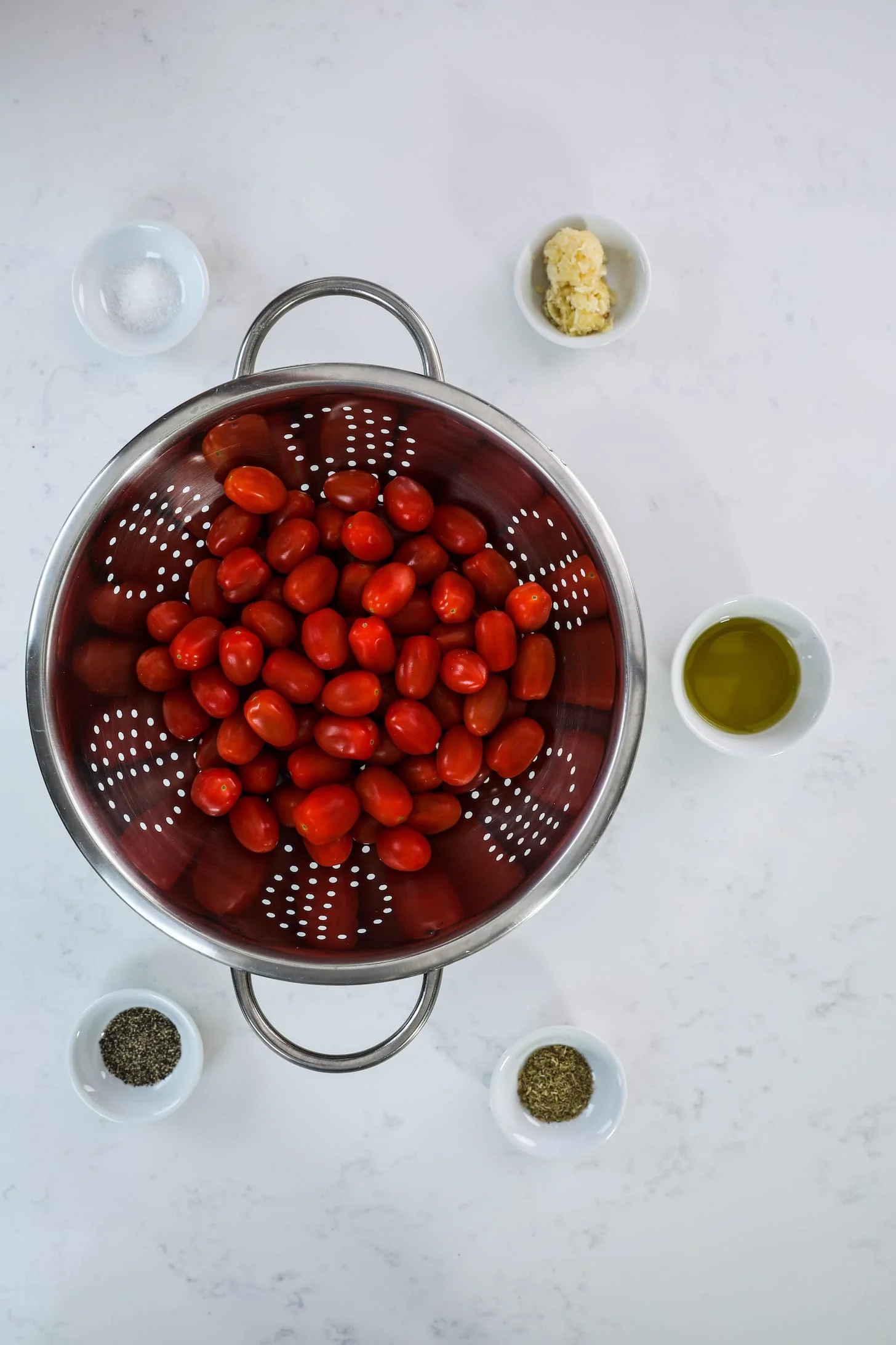 a metal sieve filled with baby red tomatoes surrounded by ramekins of ingredients, including oil, garlic, dried herbs and spices.
