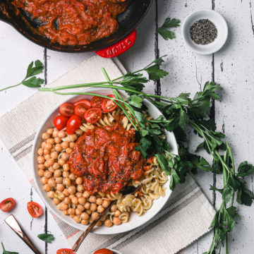 overhead shot of a bowl of pasta and chickpeas topped with thick tomato sauce styled with stems of parsley.