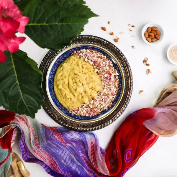 a bowl of yellow thick pudding topped with nuts and pink petals on a gold tray decorated with a multicoloured scarf on the side.