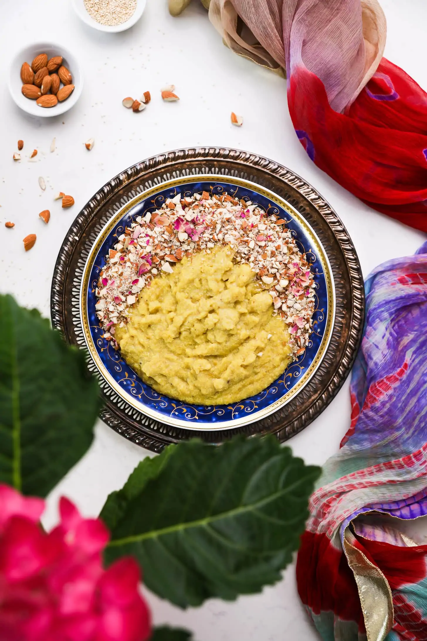 a bowl of yellow thick pudding topped with nuts and pink petals on a gold tray staged with a multicoloured scarf.