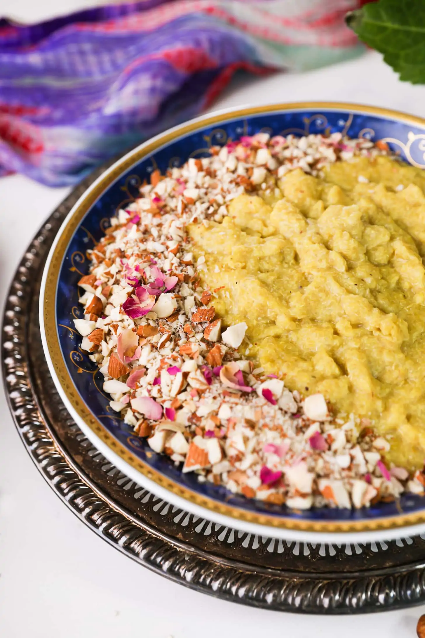 close up perspective shot of a bowl of yellow pudding topping with crushed nuts and pink petals on a gold tray.