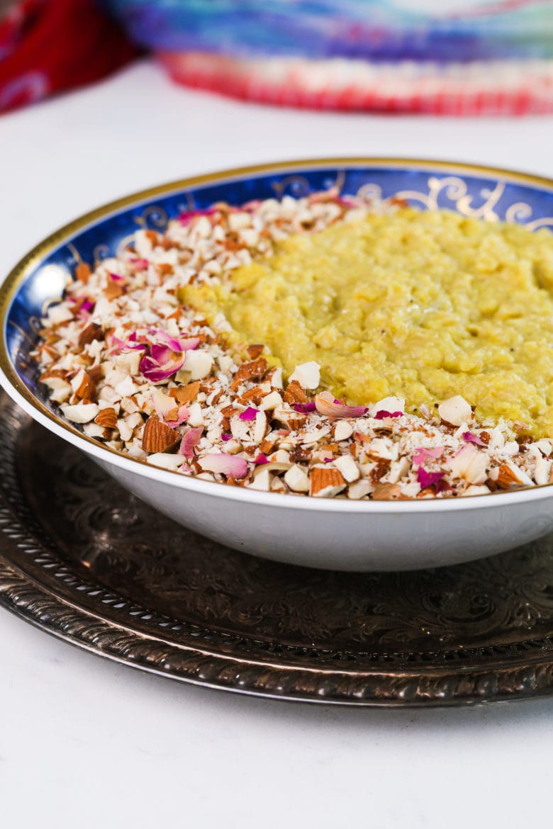 perspective shot bowl of yellow pudding topped with nuts and petals on a gold tray.
