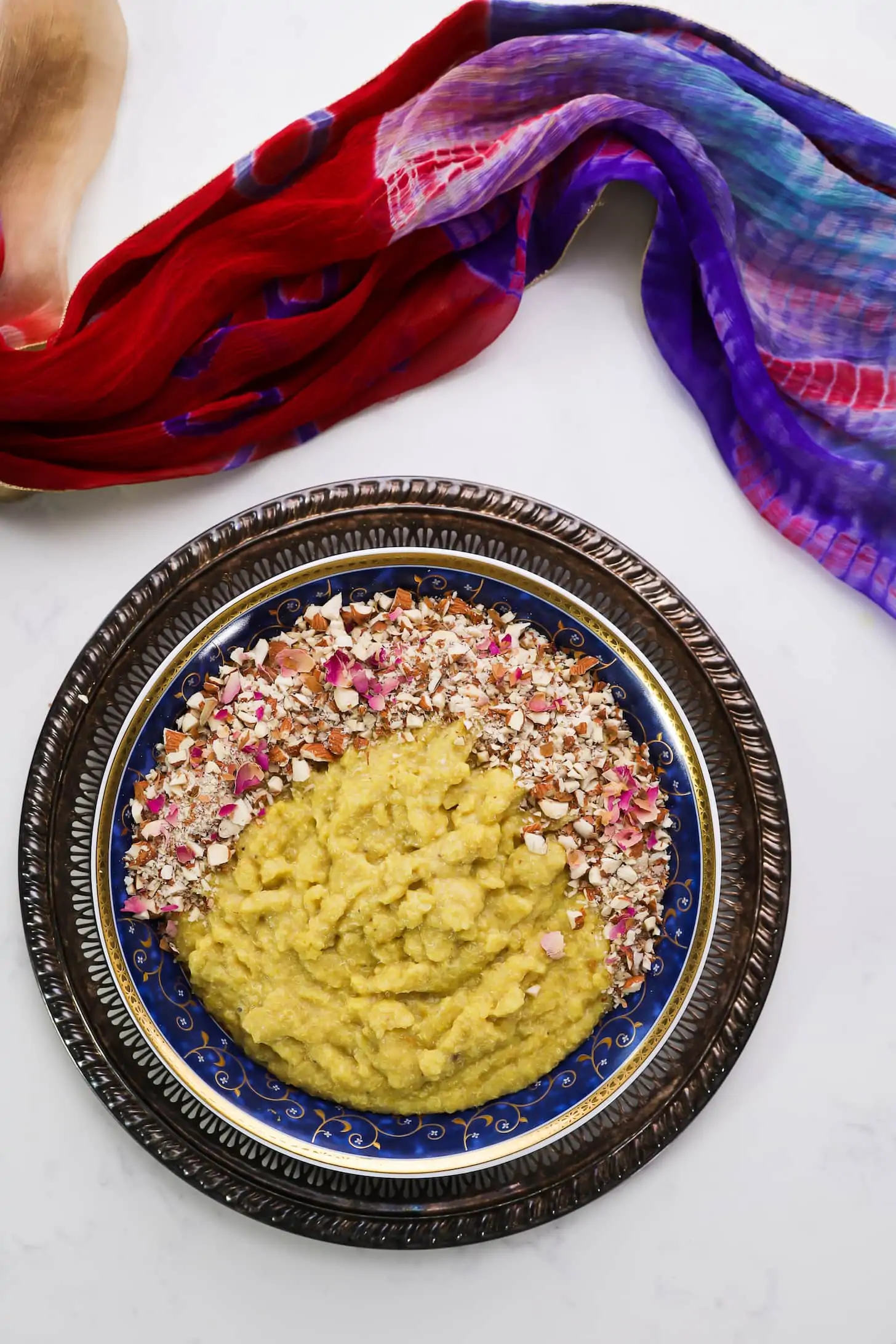 flatlay of a bowl of yellow pudding topped with nuts and pink petals on a gold tray.