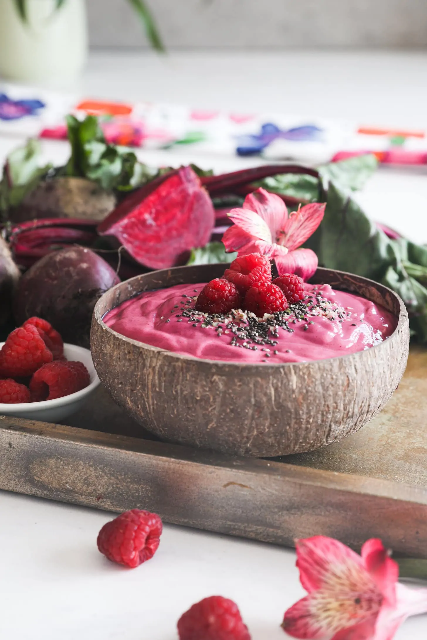 a perspective shot of a coconut bowl of a pink smoothie topped with seeds and raspberries on a gold tray styled with beets and raspberries..