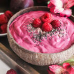 a perspective shot of a coconut bowl of a pink smoothie topped with seeds and raspberries.