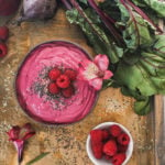 a coconut bowl of a pink smoothie topped with seeds, raspberries and a flower on a styled tray with beets and a ramekin of raspberries.