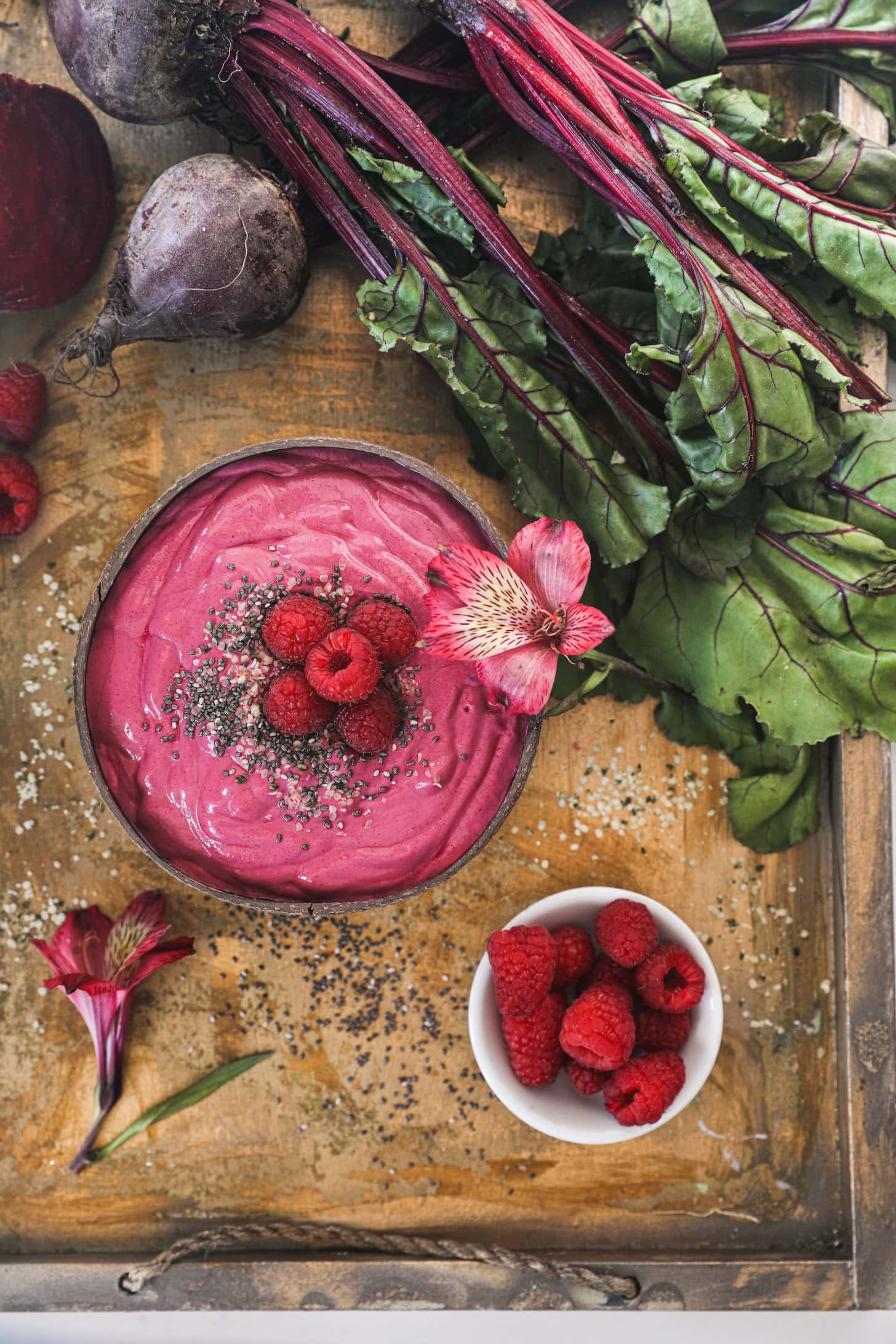 a coconut bowl of a pink smoothie topped with seeds, raspberries and a flower on a styled tray with beets and a ramekin of raspberries.