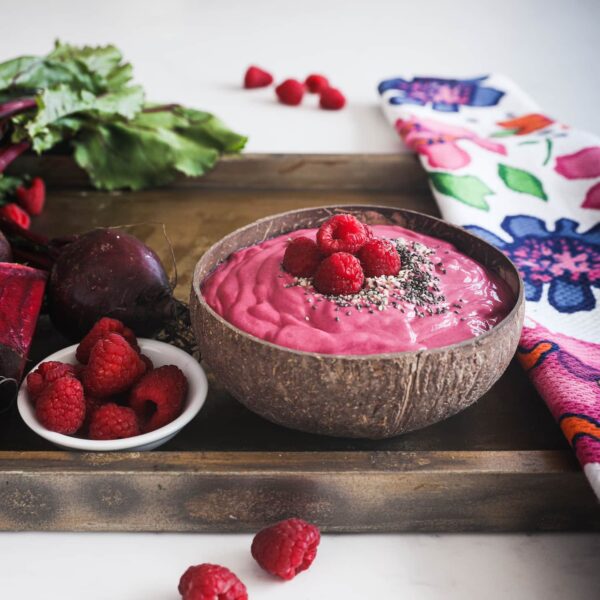 a coconut bowl of a pink smoothie topped with seeds and raspberries on a styled tray