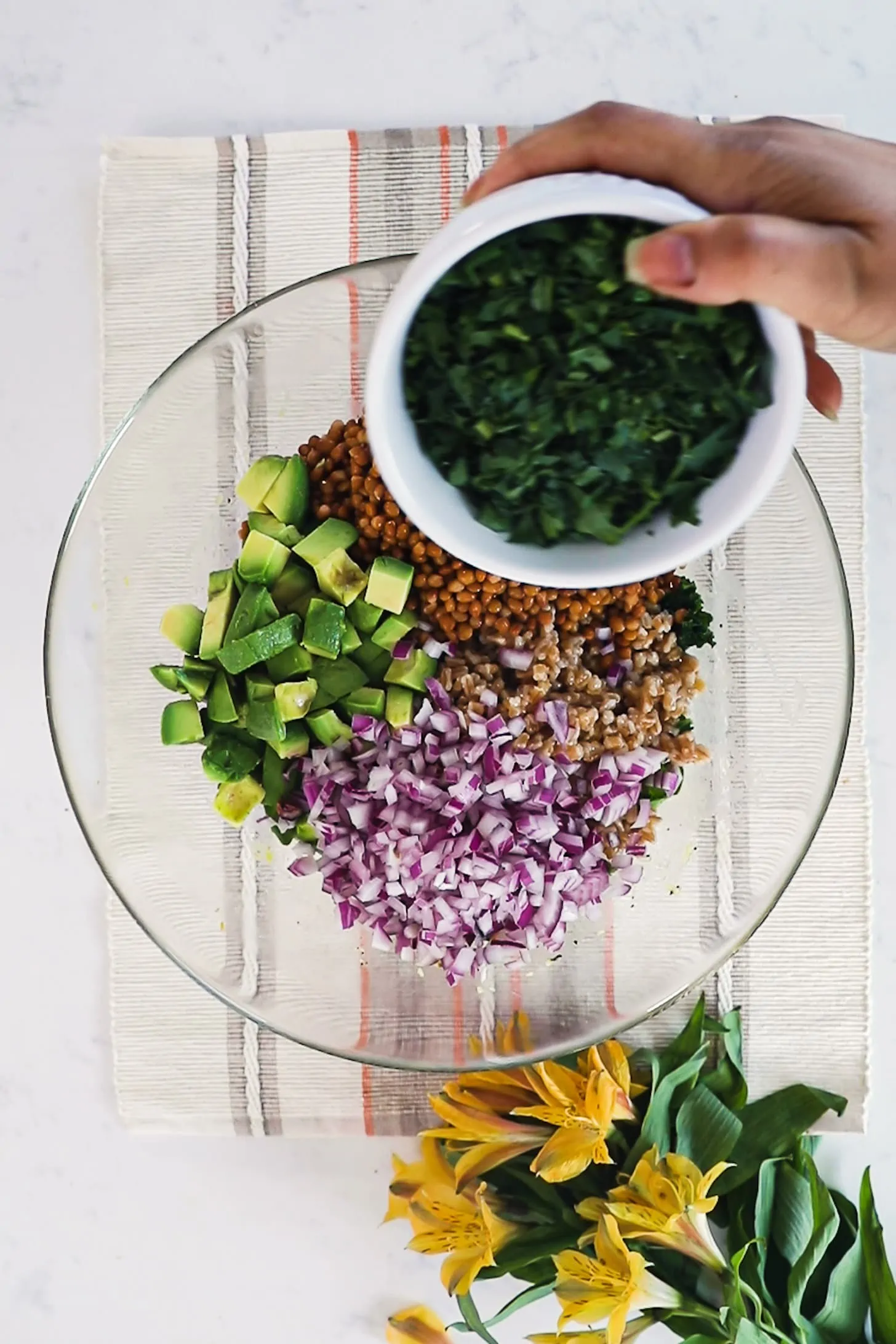a hand holding a bowl of greens over a bowl of chopped onions, avocados and lentils.