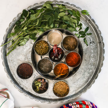 a silver round tray with a spice box containing small ramekins of dry and powdered spices and pile of green leaves on stems