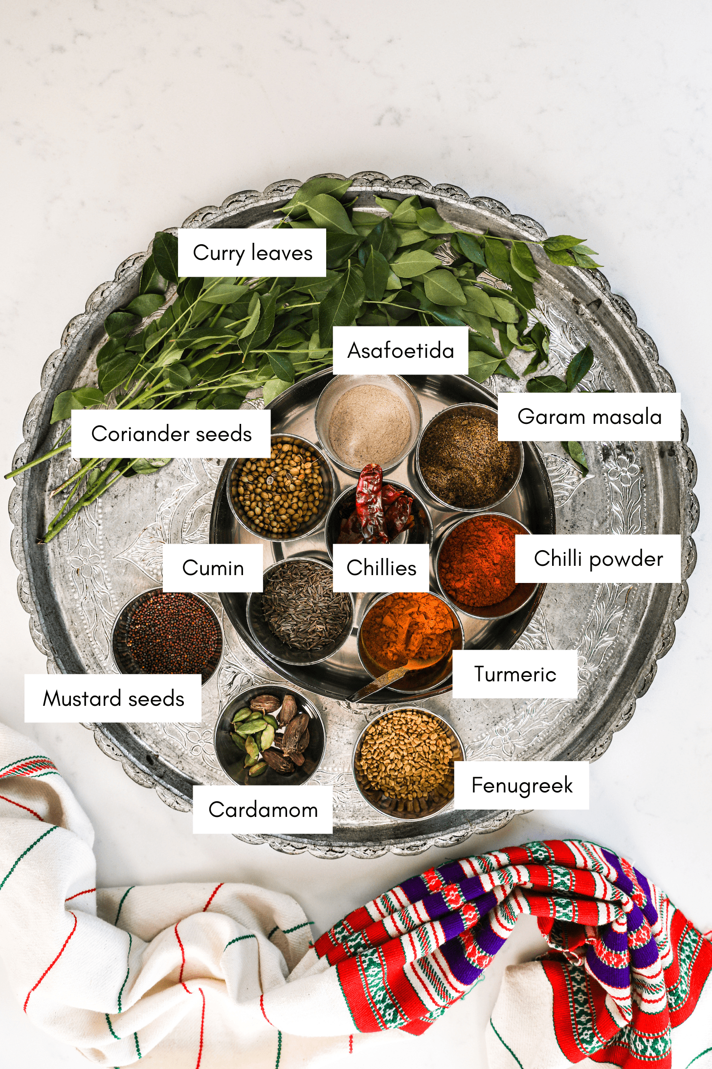a silver round tray with a spice box containing small ramekins of dry and powdered spices and pile of green leaves on stems and a traditional South Asian scarf styles on the side.