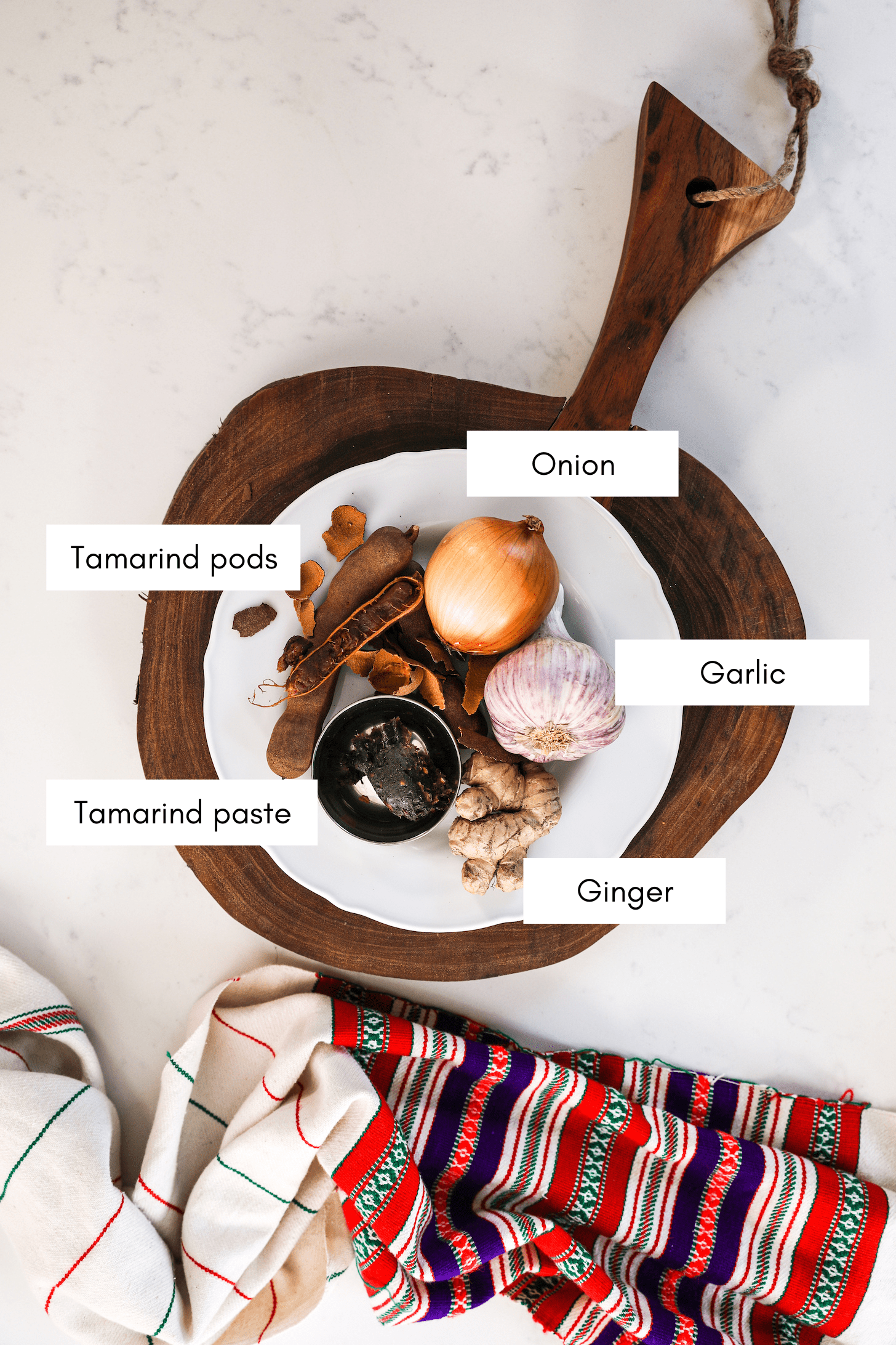 a wooden board with a plate of aromatics including onion, garlic, ginger and tamarind in different forms.