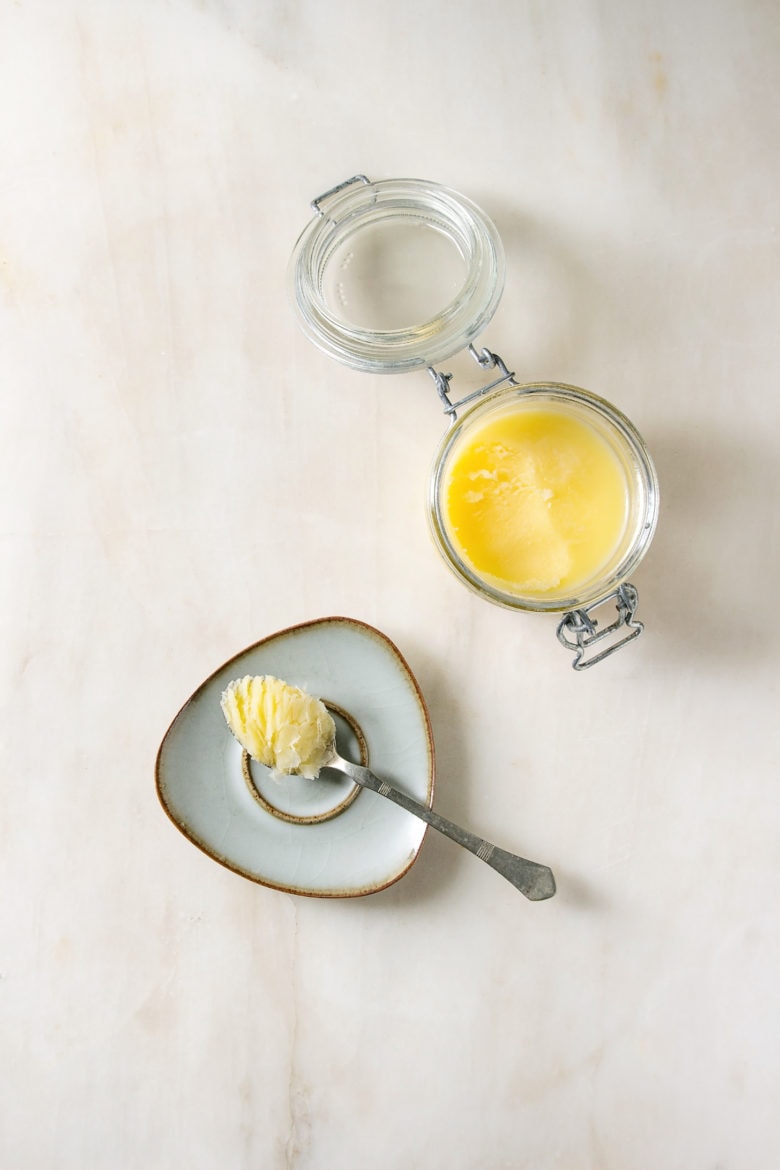 Homemade Melted ghee clarified butter in open glass jar and spoon on saucer over white marble background. Flat lay, space