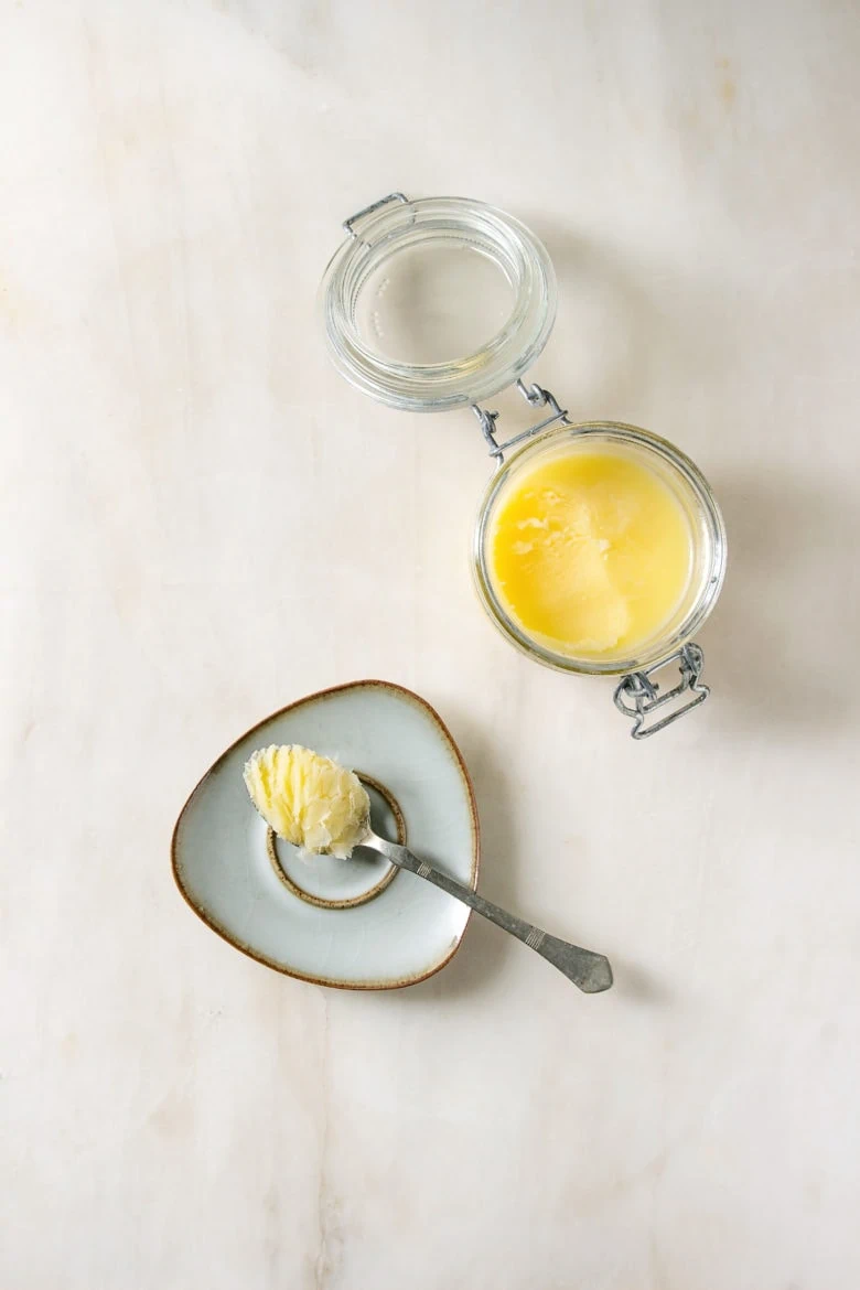Homemade Melted ghee clarified butter in open glass jar and spoon on saucer over white marble background. Flat lay, space