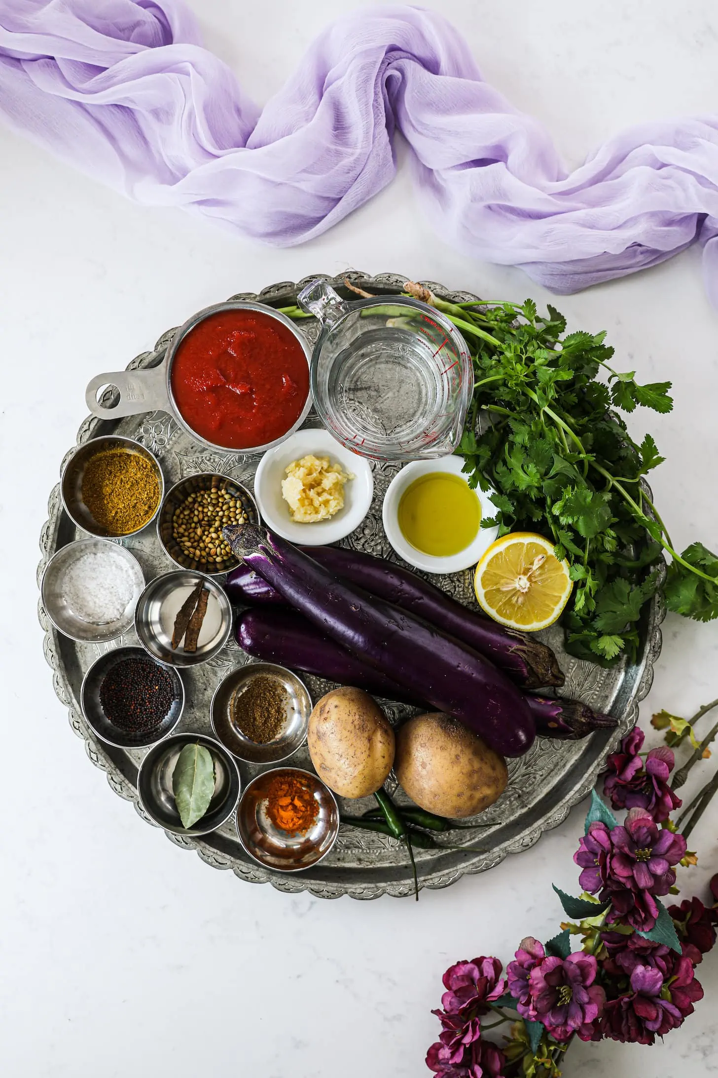 a silver round tray with an array of spices and food ingredients styled with a lilac scarf and flowers.