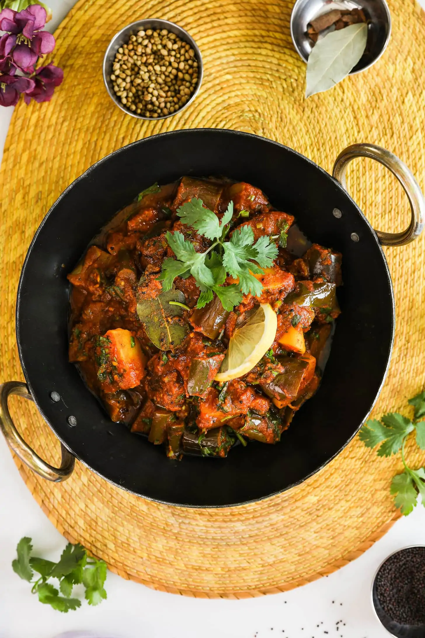 wok of cooked vegetables topped with a lemon slice and cilantro leaves on a yellow place mat styled around ramekins of spices.