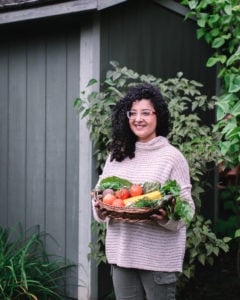 a lady standing in front of a garden shed holding a basket of vegetables.