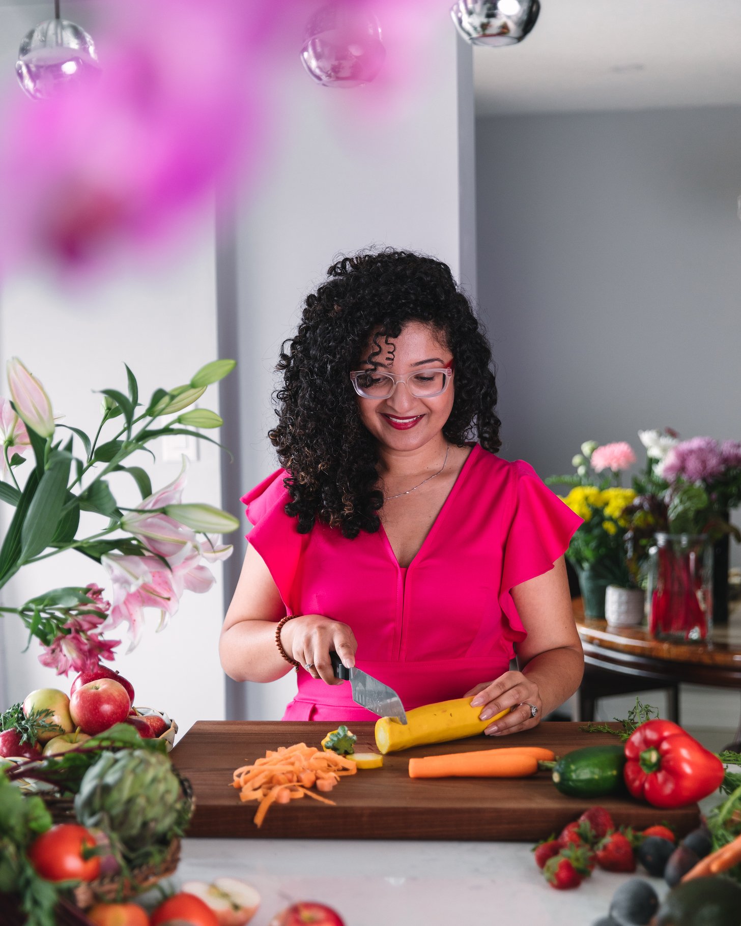 a curly haired lady in her kitchen chopping a yellow vegetable surrounded by food and flowers.