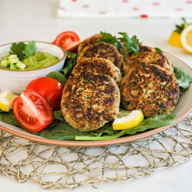 close up of a plate of cooked patties on a bed of greens with tomatoes and lemon slices arranged around them.