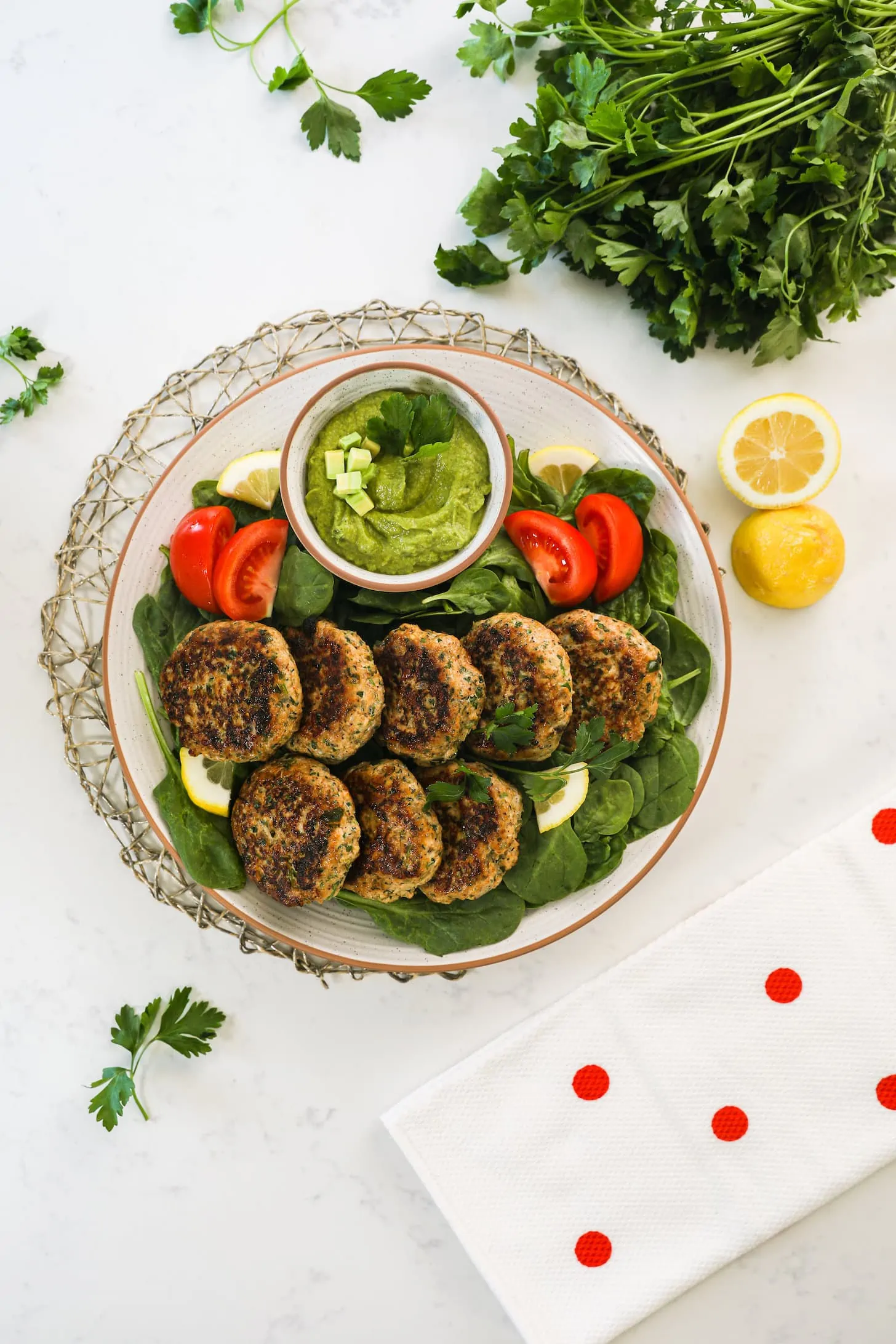 Flat lay shot of a plate of cooked patties on a bed of greens with tomatoes and lemon slices arranged around them and a bowl of green dip on the side.