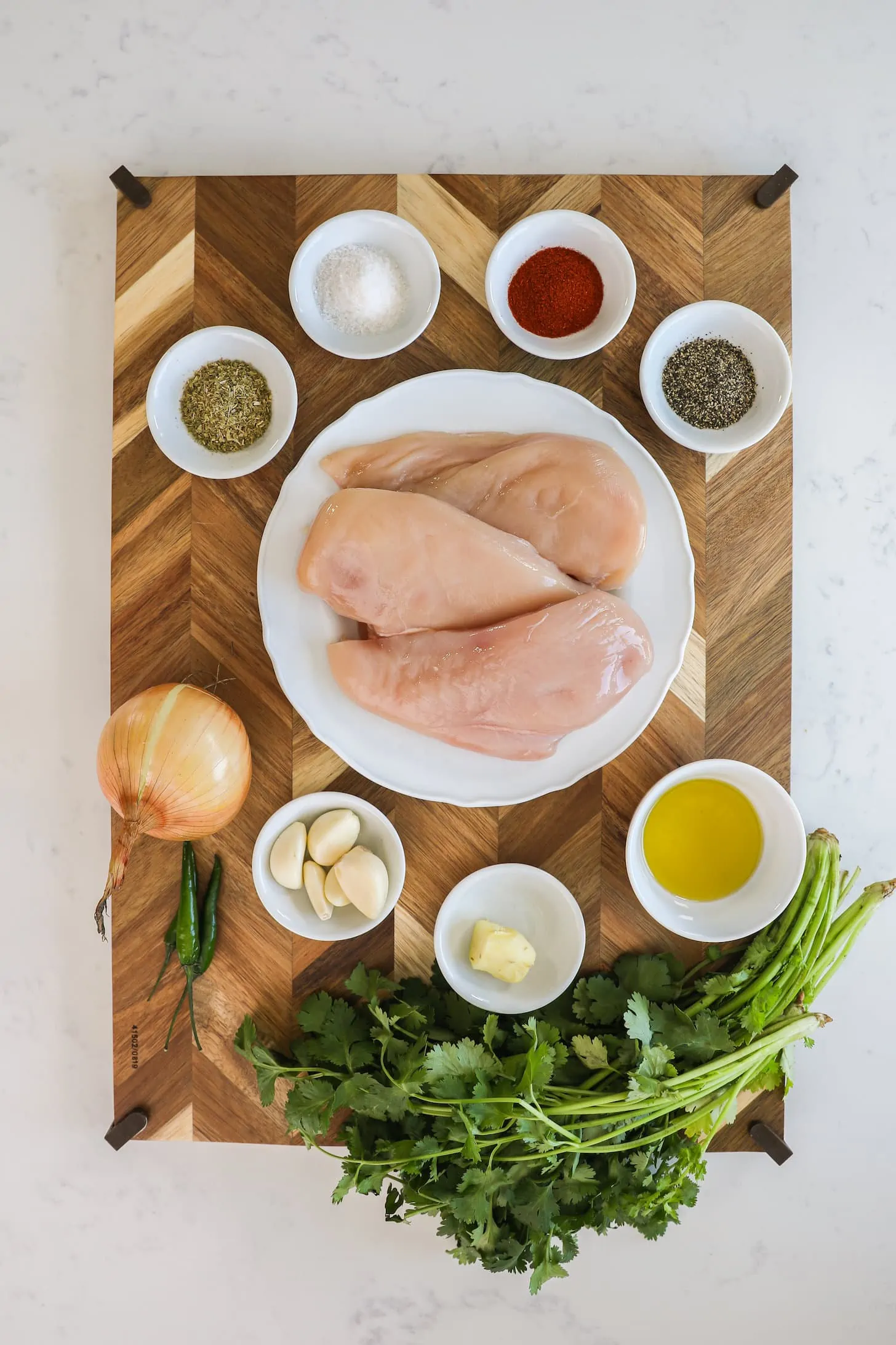 a wooden board with food ingredients arranged on it including raw chicken breast, herbs, spices and oil.