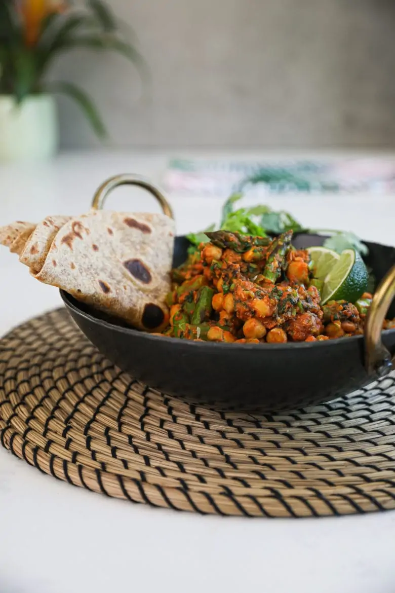 perspective shot of a wok filled with cooked chickpeas and asparagus in an orange/red sauce with a side of folded chapati topped with lime segments. There is also a plant in the background.