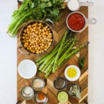 a wooden board staged with food ingredients like asparagus, a wok of chickpeas, spices, herbs and chillies.