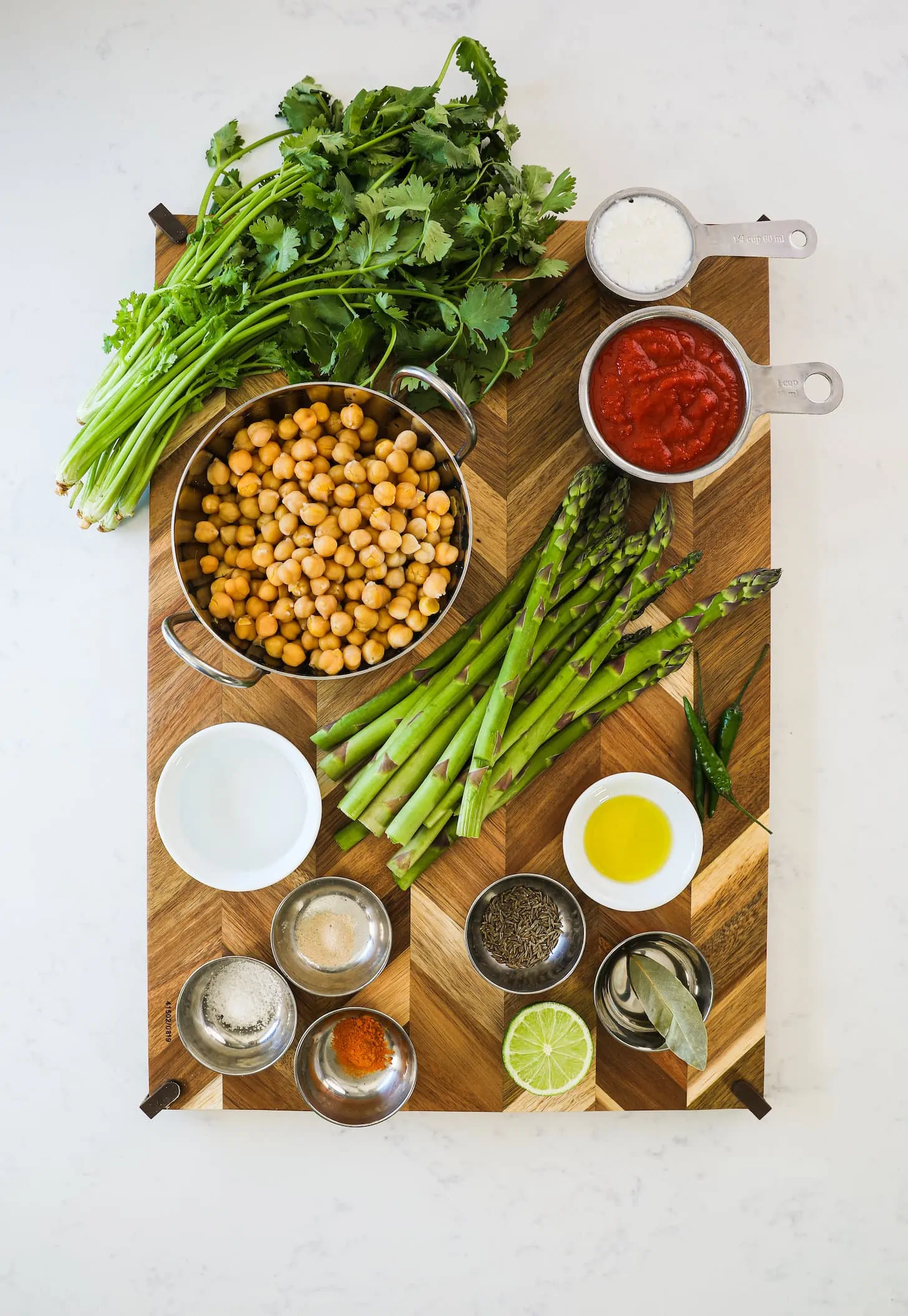 a wooden board staged with food ingredients like asparagus, a wok of chickpeas, spices, herbs and chillies.