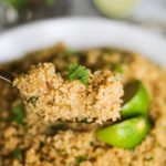 a close up shot of a fork of cooked quinoa with a bowl of cooked quinoa in the background.