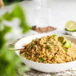 a bowl of cooked yellow quinoa topped with lime wedges and cilantro leaves. There are some greens blurred in the foreground and a hand holding a bottle of water and pouring it into a glass.