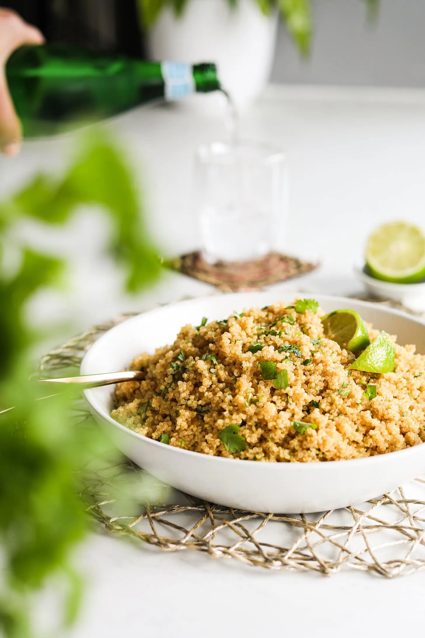 a bowl of cooked yellow quinoa topped with lime wedges and cilantro leaves. There are some greens blurred in the foreground and a hand holding a bottle of water and pouring it into a glass.