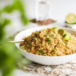 a bowl of cooked yellow quinoa topped with lime wedges and cilantro leaves. There are some greens blurred in the foreground and a glass of water in the background.