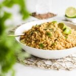 a bowl of cooked yellow quinoa topped with lime wedges and cilantro leaves. There are some greens blurred in the foreground.