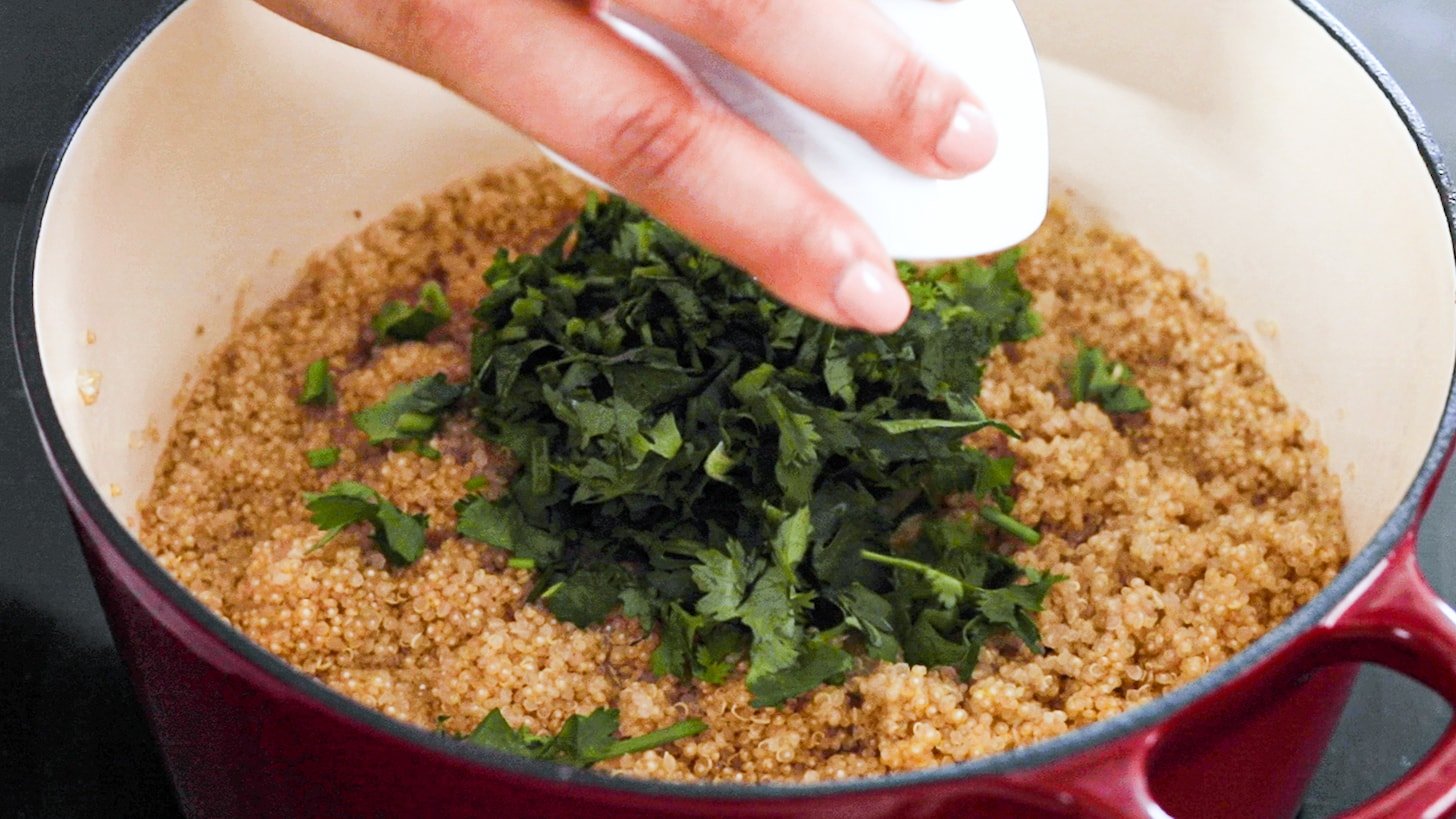 a hand holding a ramekin of cilantro being added to a pot of quinoa.