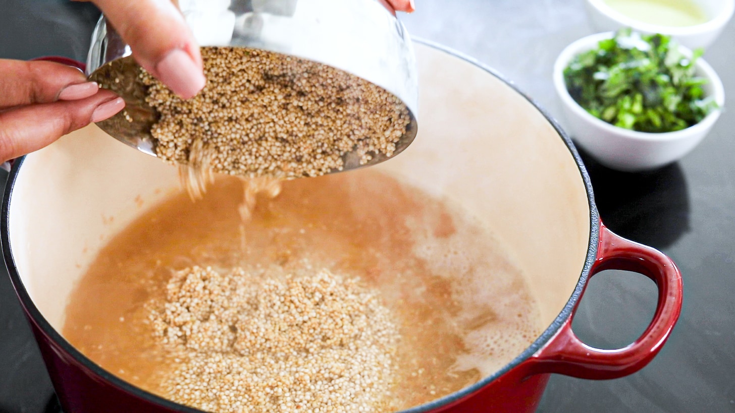 a hand holding a bowl of quinoa that is being added to a pot of quinoa and stock..