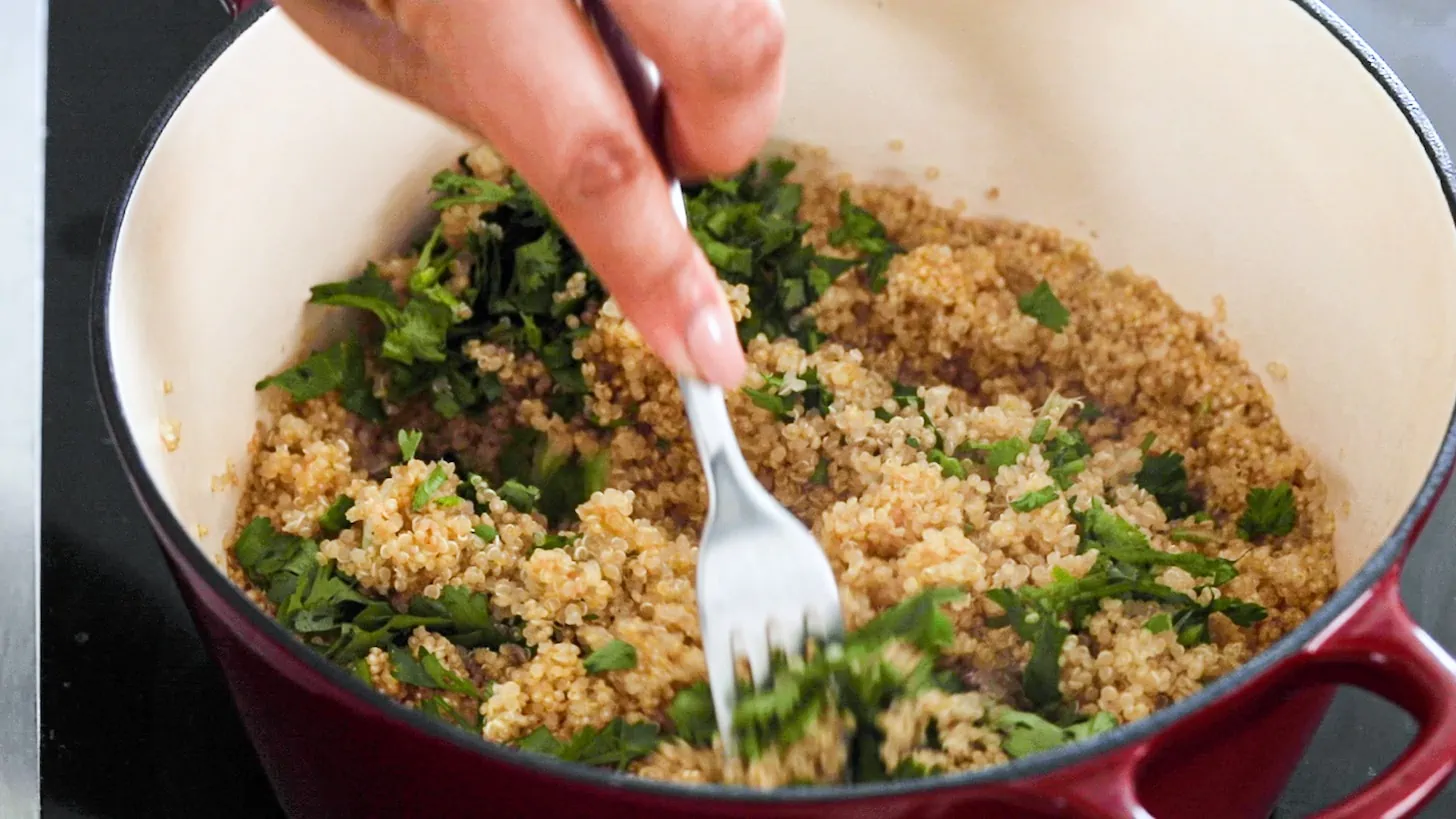 a hand holding a fork fluffing cooked quinoa with chopped cilantro.