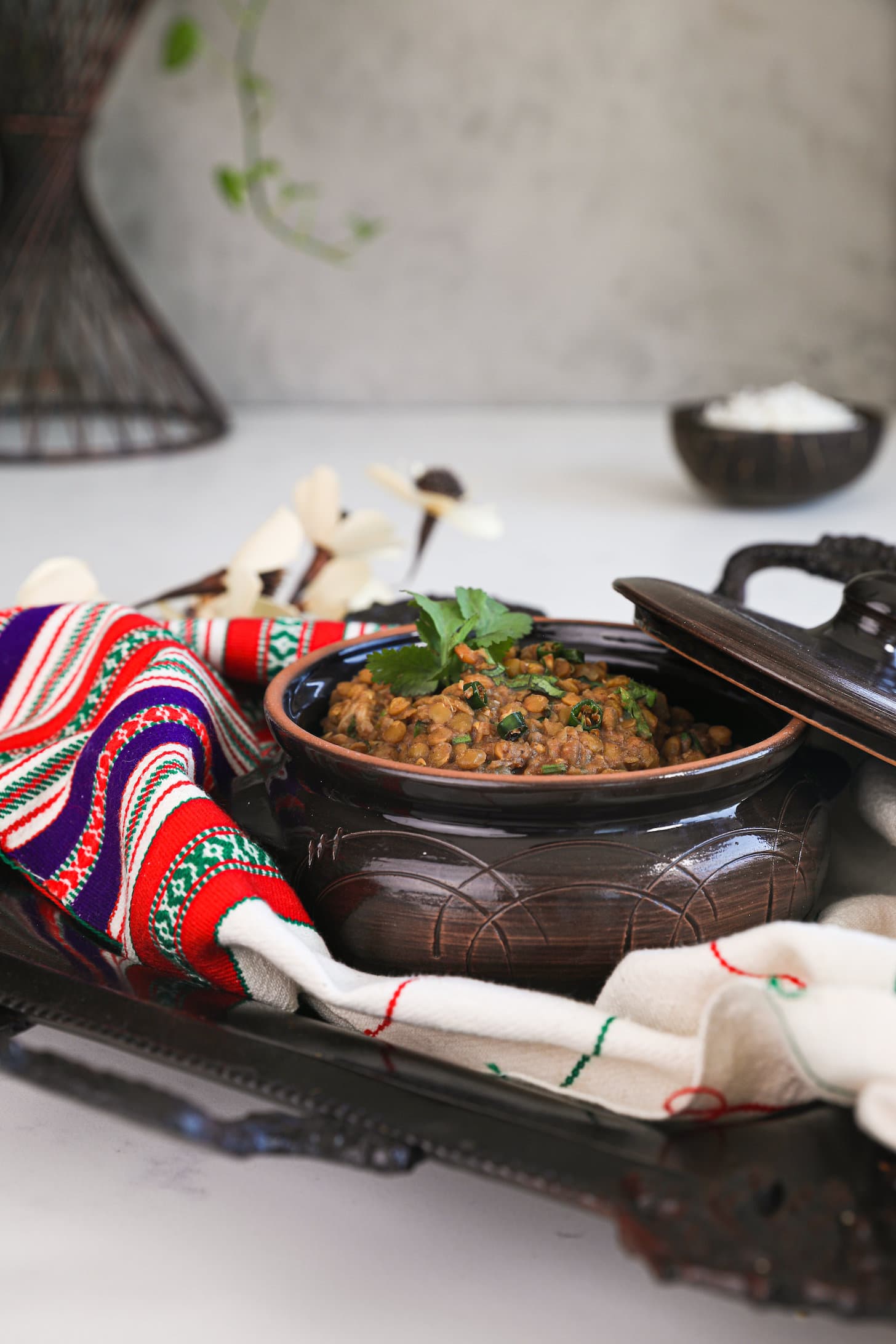 perspective image of a round clay container with cooked lentils in a tray styled with a cream coloured scarf. There is a bowl of rice in the background.