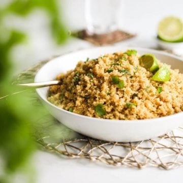 a bowl of cooked yellow quinoa topped with lime wedges and cilantro leaves. There are some greens blurred in the foreground and a glass of water in the background.