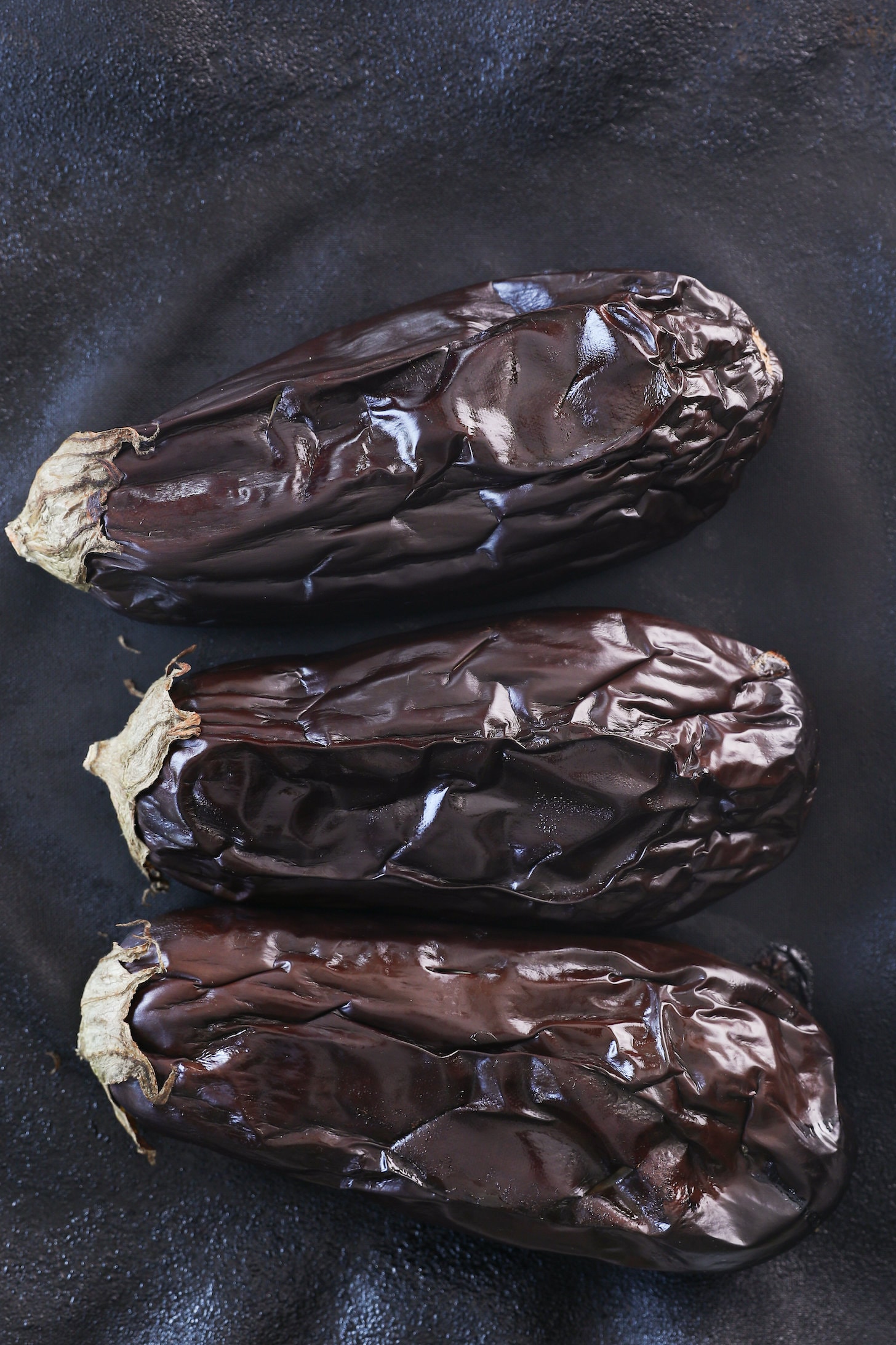 Three cooked eggplants side by side on a black plate.