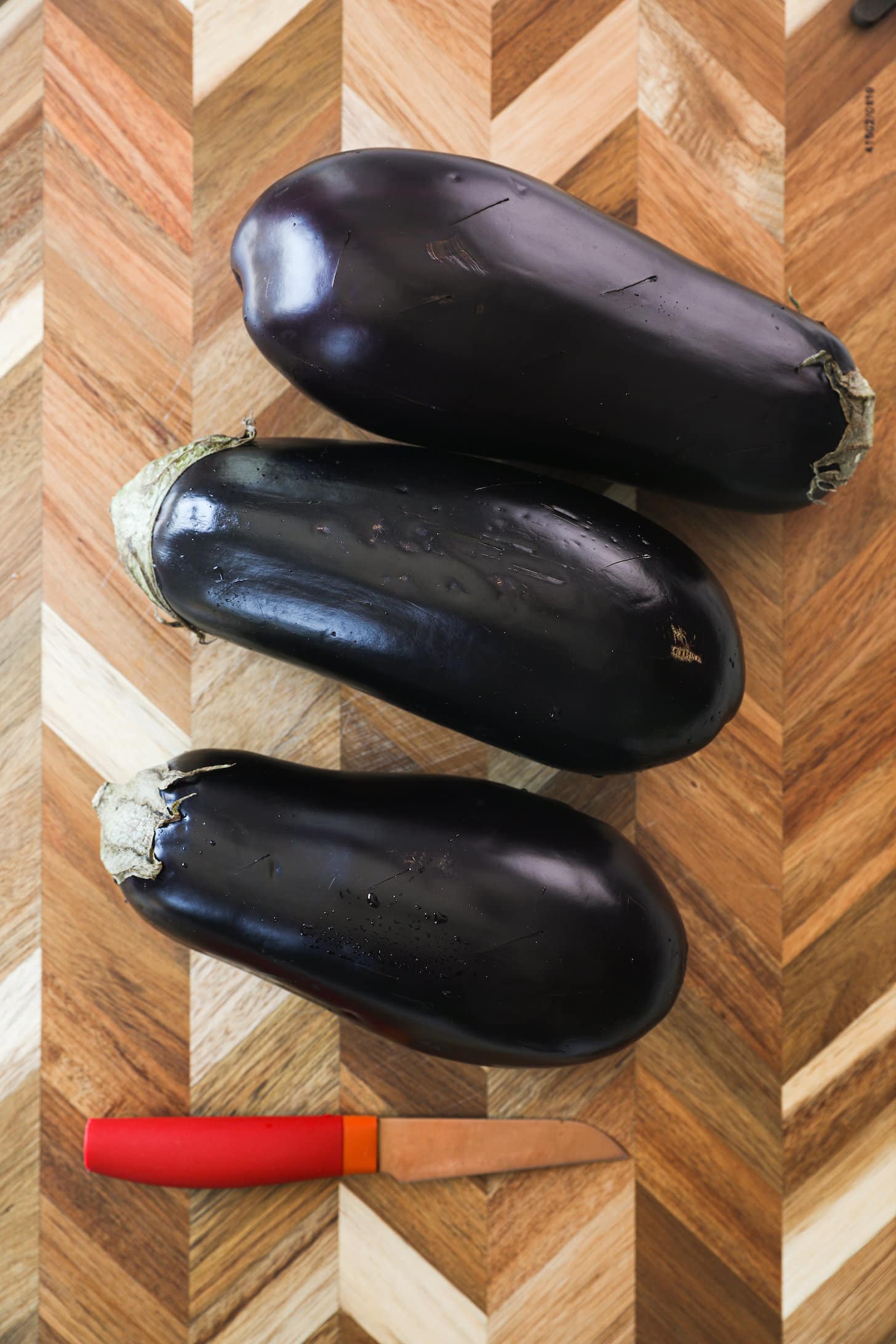 Three globe eggplants on a wooden board with a red knife next to them.