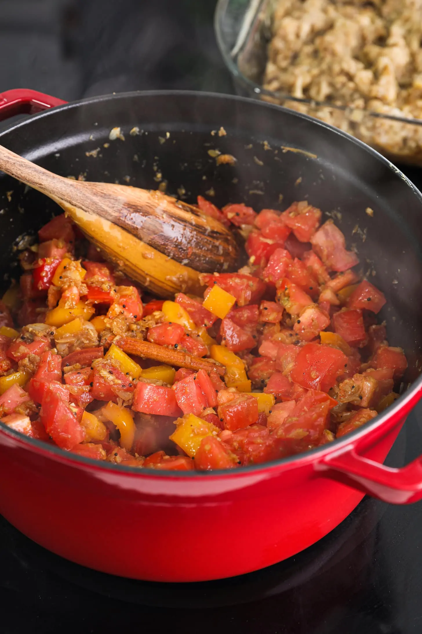 a red cooking pot full of chopped tomatoes and orange peppers with a wooden spoon immersed in it.