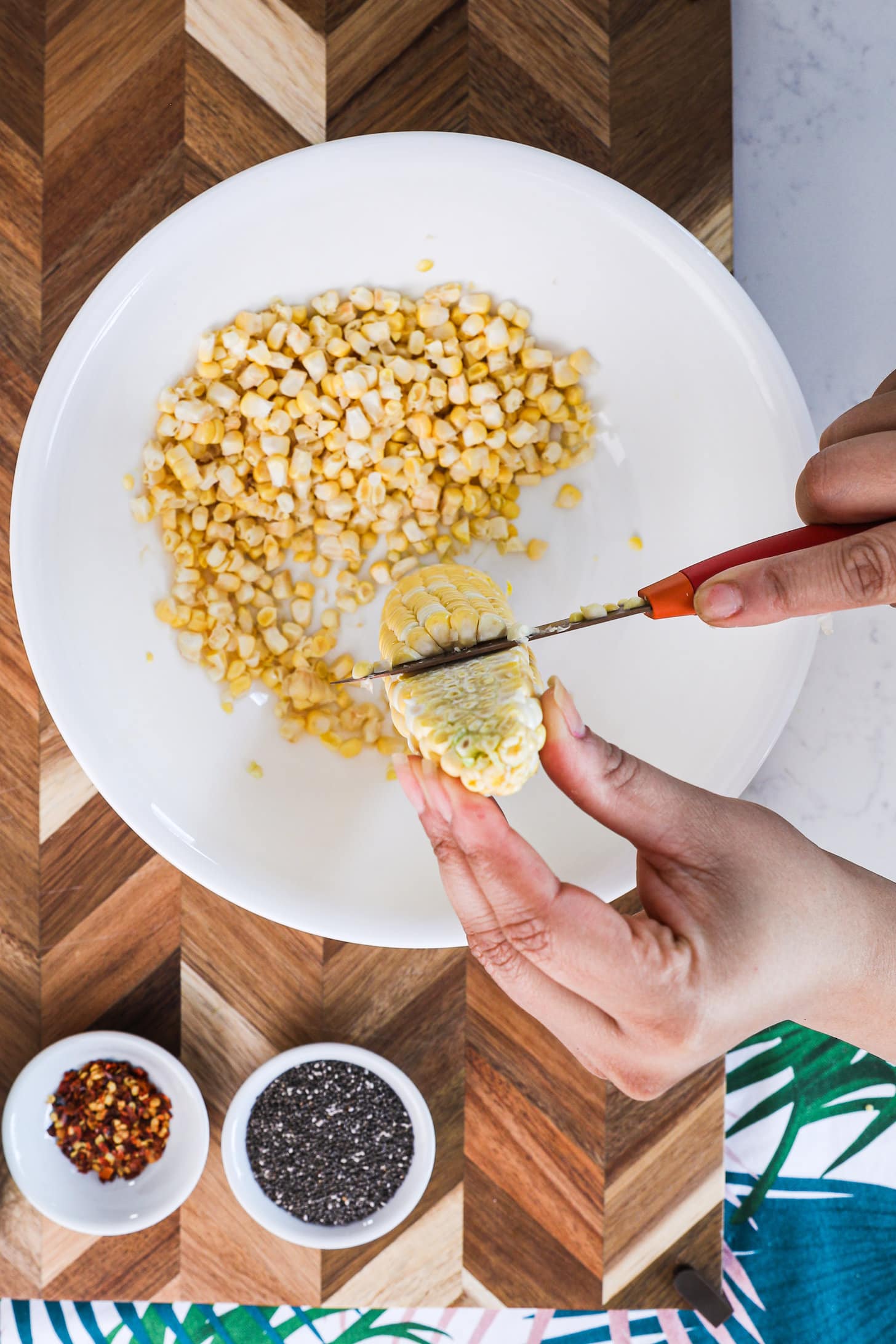 a hand holding a knife and cutting the kernels off of a cob of corn into a white bow.