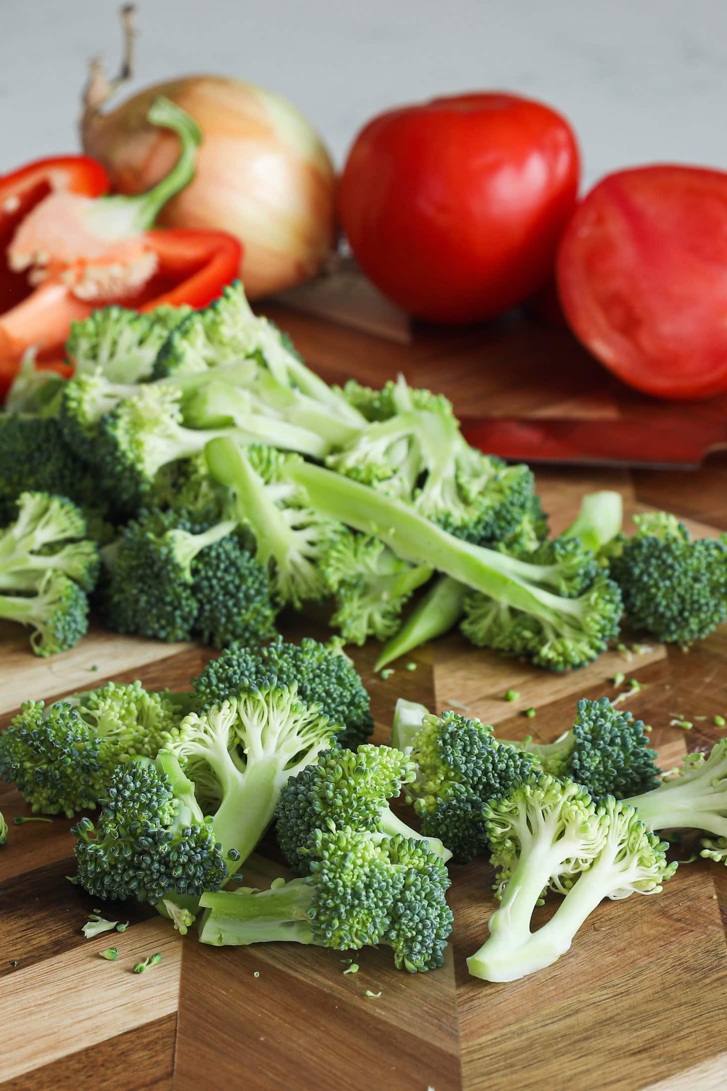 Chopped florets of broccoli on a wooden board with other vegetables in the background.