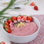 Perspective image of a pink smoothie topped with strawberry halves, lemon segments and pink flowers. It's placed on a pink mat with ramekins of seeds in the background.