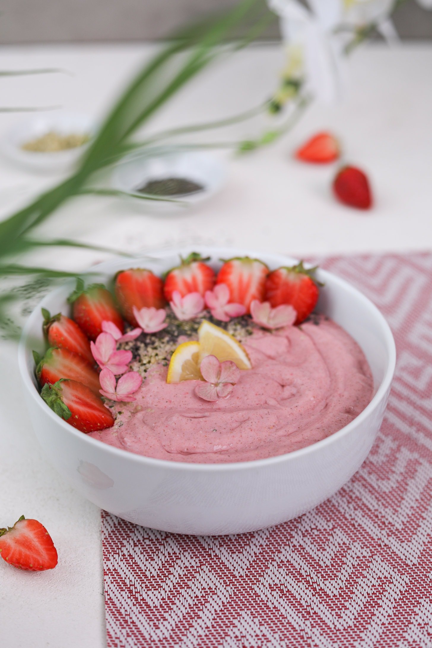 Perspective image of a pink smoothie topped with strawberry halves, lemon segments and pink flowers. It's placed on a pink mat with ramekins of seeds in the background.