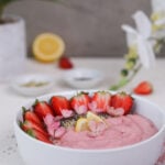 Perspective image of a pink smoothie topped with strawberry halves, lemon segments and pink flowers. It's placed on a pink mat with ramekins of seeds and plants in the background.